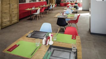 A modern restaurant with wooden tables and colorful chairs, a red booth seating area, and a kitchen counter in the background.