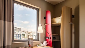 A compact hotel room with a desk, chair, and a bed with a red headboard. A window offers a city view, and a lamp is on the desk. - ibis Istanbul West (Istanbul)