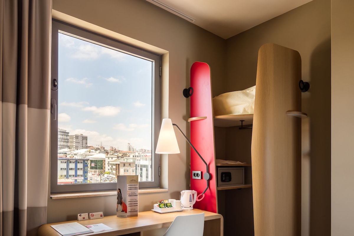 A compact hotel room with a desk, chair, and a bed with a red headboard. A window offers a city view, and a lamp is on the desk.