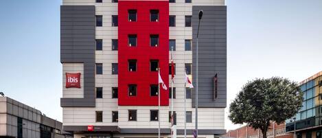 A modern hotel building with a prominent red and grey facade, multiple windows, and a visible entrance.