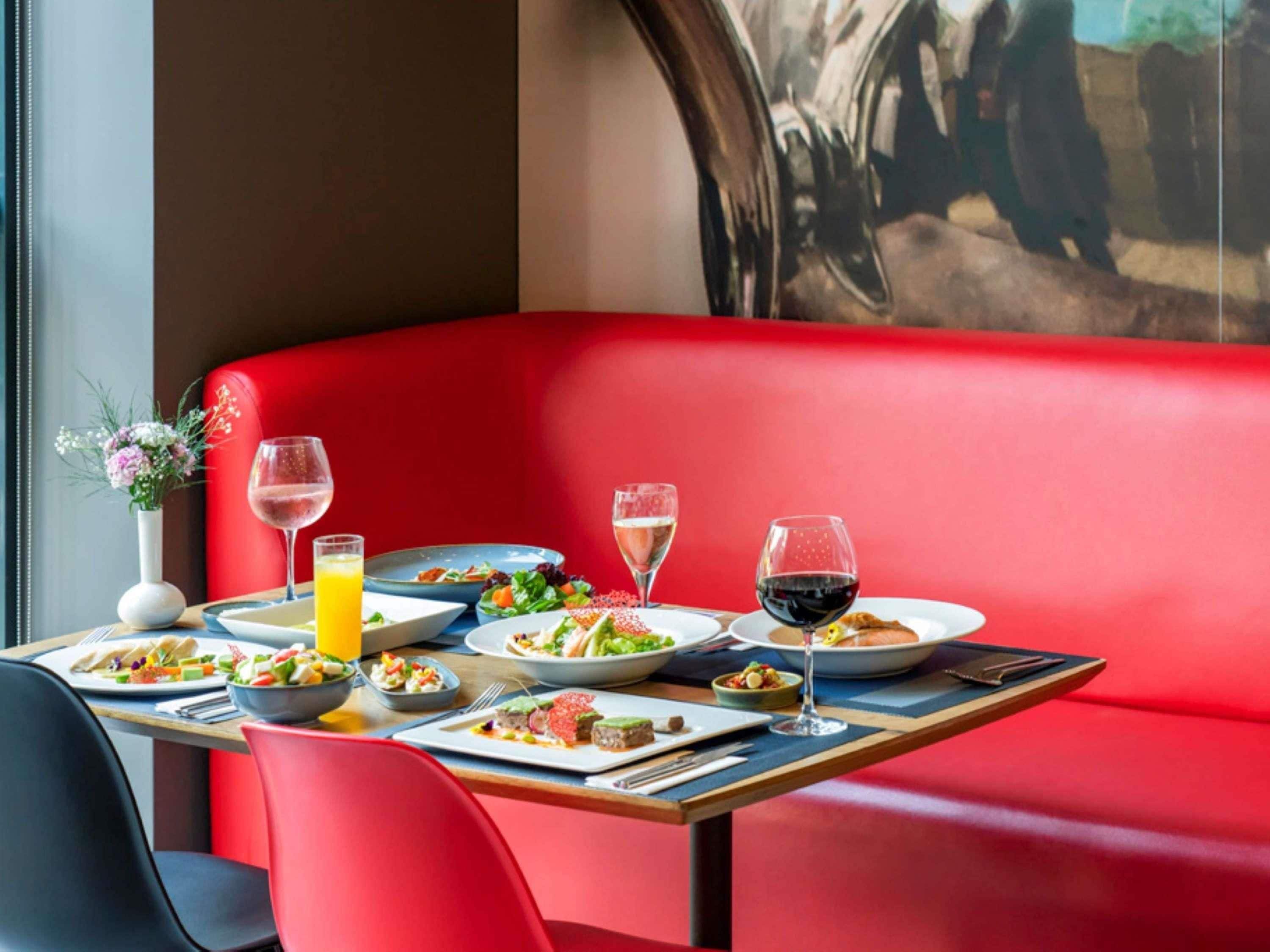 Une table à manger, dressée avec divers plats et boissons, est placée devant un banc recouvert d’un tissu rouge.