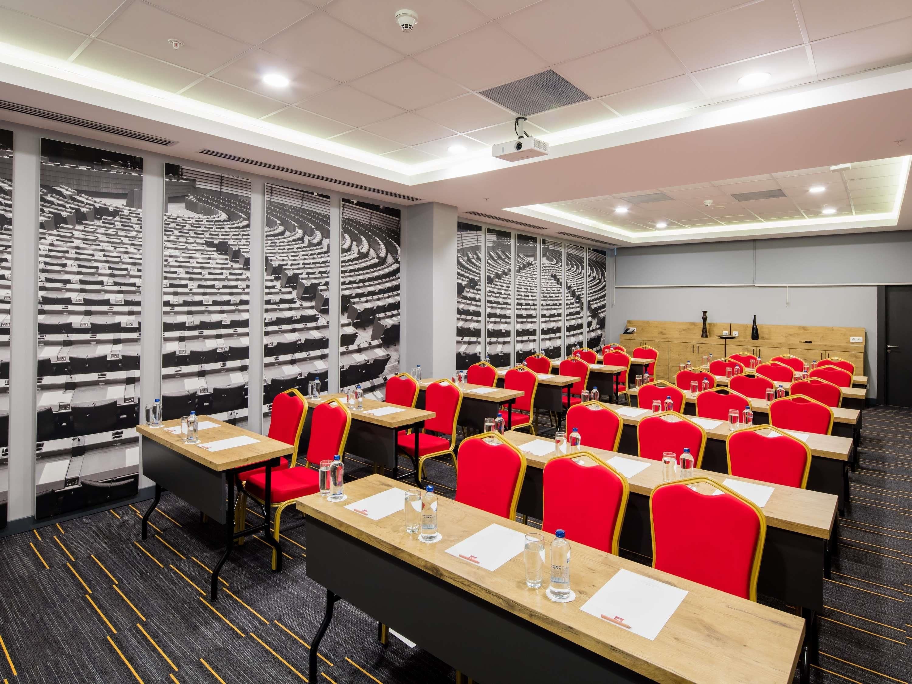 A conference room with rows of red chairs, wooden tables, and a large screen wall.