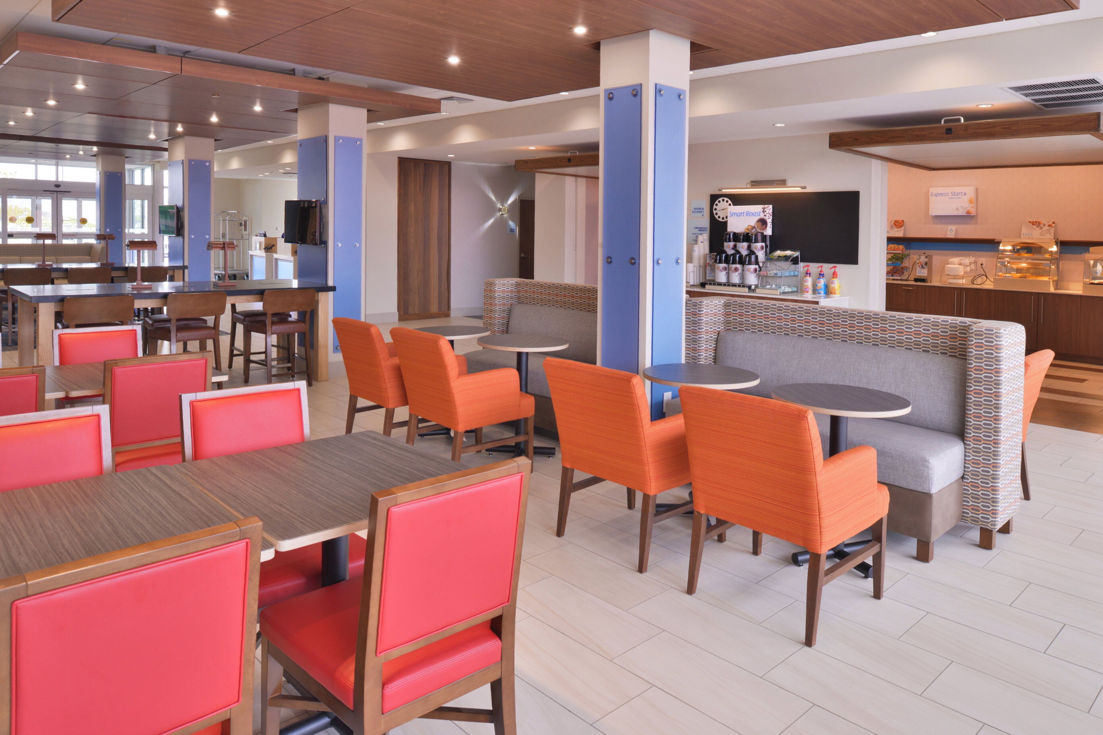 A modern cafe with wooden tables and chairs, a bar area, and a seating section with patterned cushions.