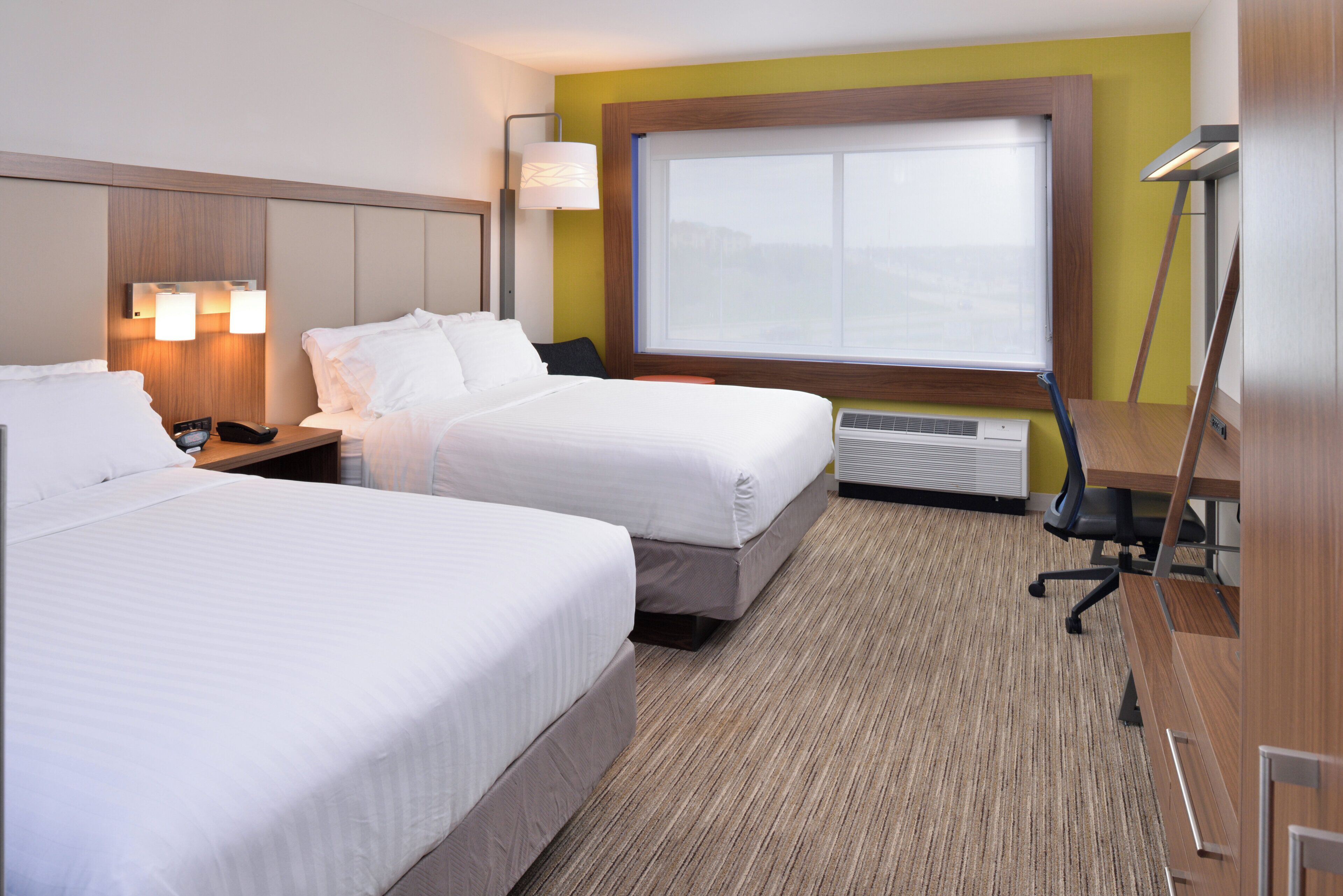 A hotel room with two beds, a desk, a chair, a lamp, and a large window.