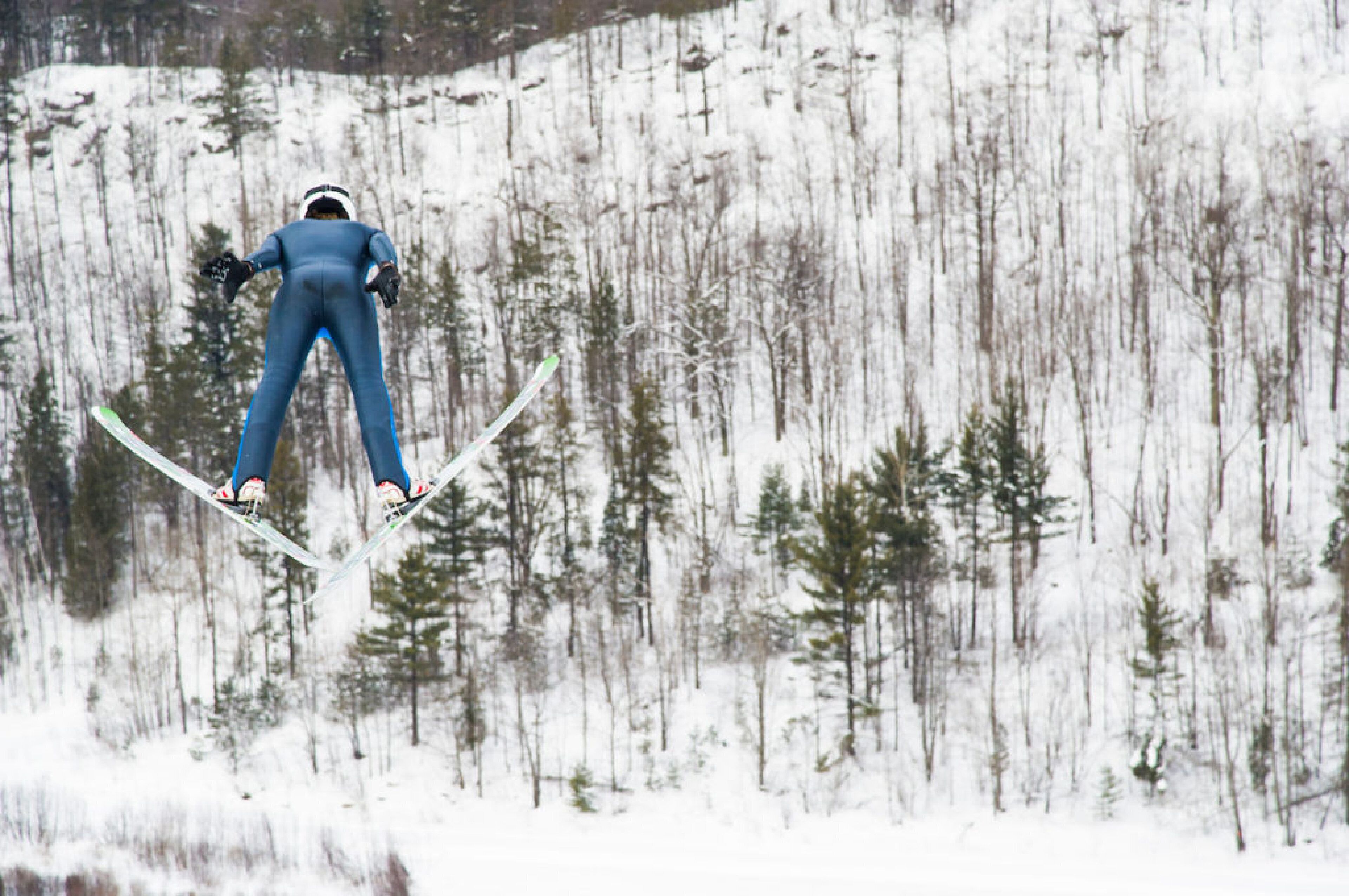 A skier in mid-air performing a jump with skis, surrounded by a snowy forest.