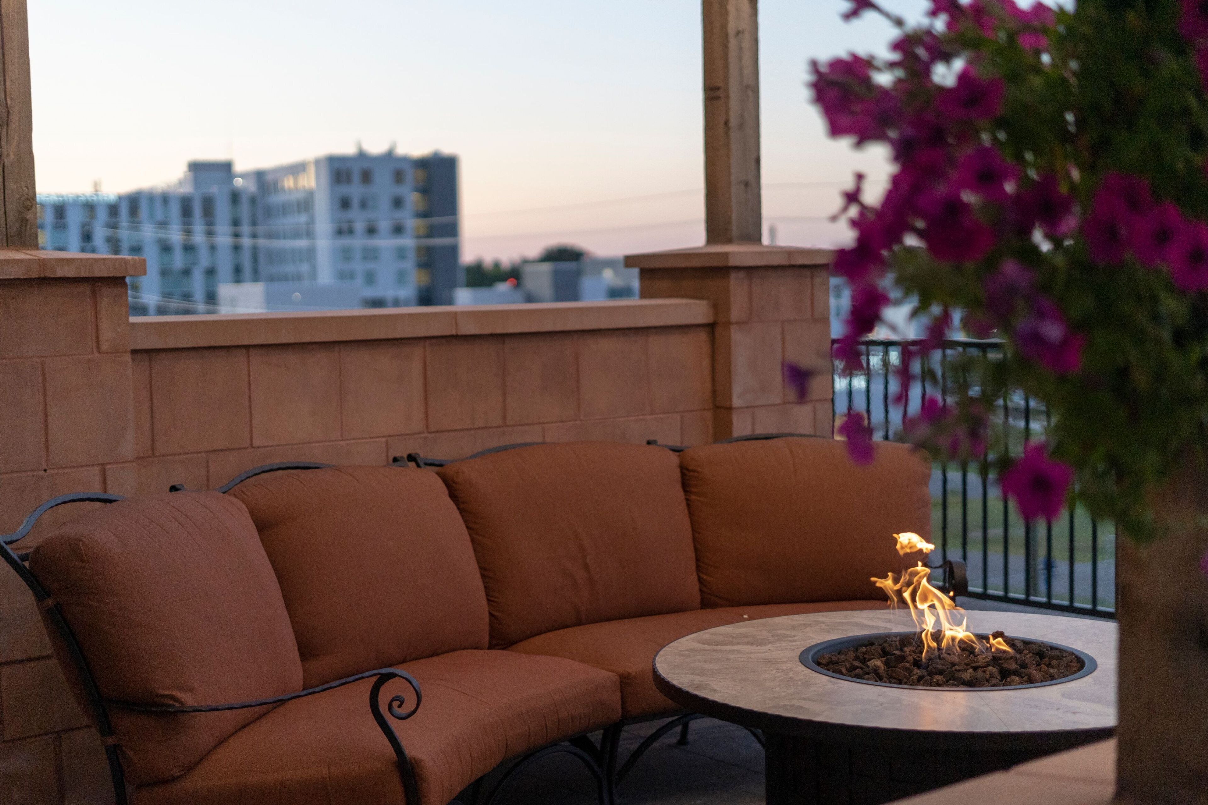 A section of a building with a curved outdoor seating area, a fire pit, and a view of buildings in the background.