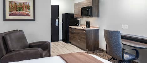 A hotel room with a bed, a desk, a chair, a kitchenette, and a framed picture on the wall.