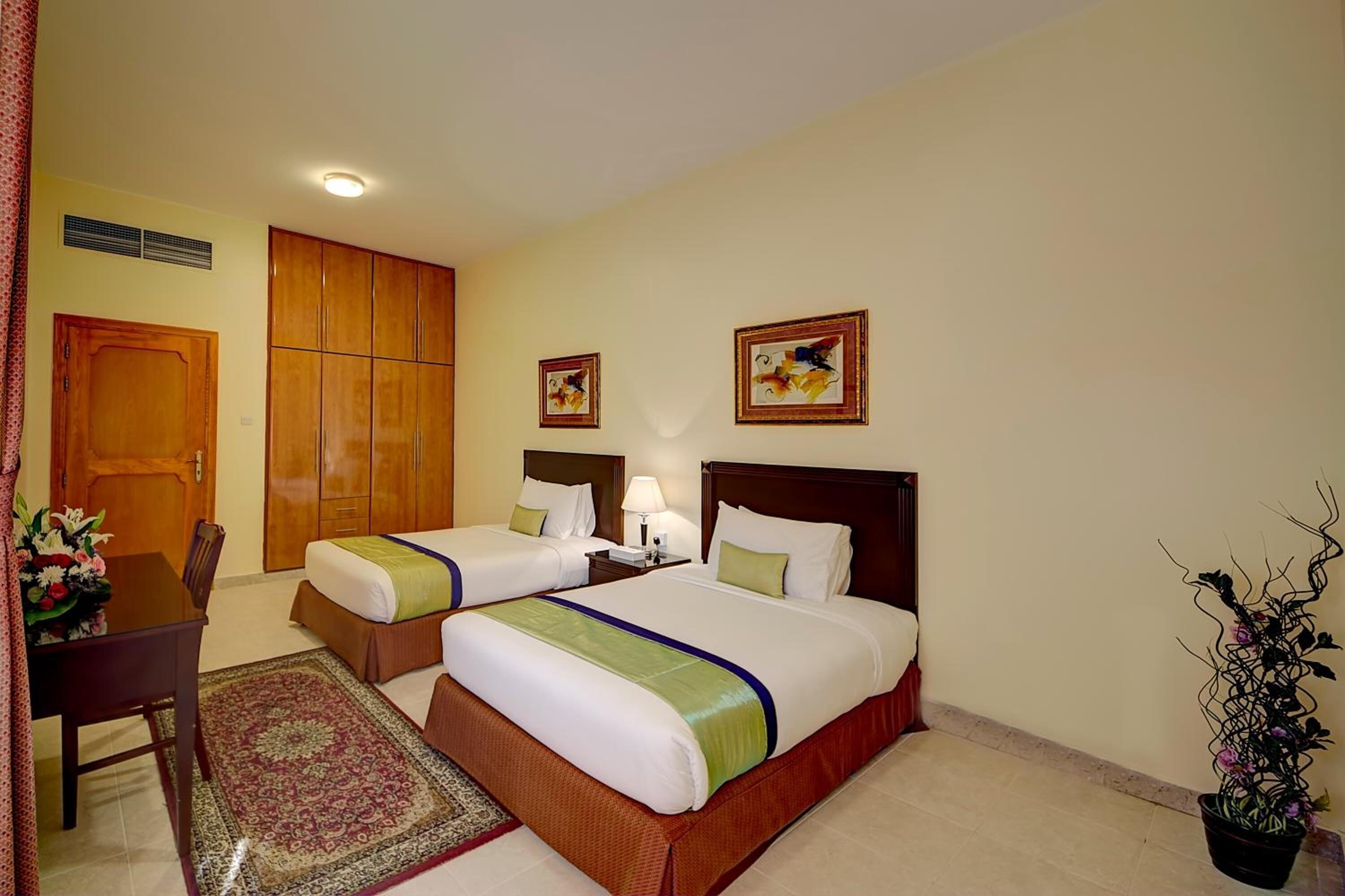 A hotel room with two beds, a desk, a chair, a dining table, and a potted plant.