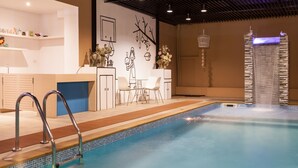 Une piscine intérieure avec un bord carrelé, un espace détente avec des chaises blanches, un mur décoratif orné d’une branche d’arbre, et une structure haute et étroite surmontée d’oiseaux.