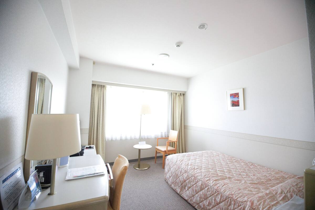 A hotel room with a bed, a desk with a lamp, a chair, a small table, and a window with curtains.