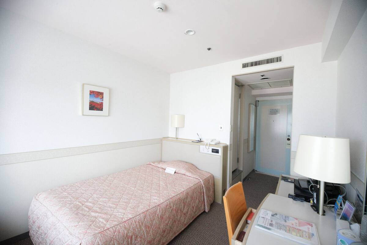 A hotel room with a single bed, a desk with a lamp, a chair, and a door leading to another room.