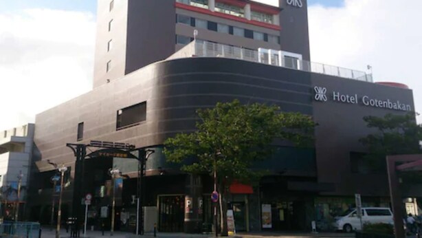 Un hotel moderno con un llamativo letrero que dice 'Hotel Gotenbakan', un amplio toldo de entrada y un estacionamiento visible al frente.