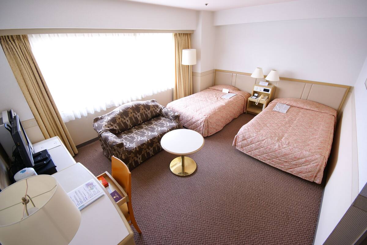 A hotel room with a sofa, two single beds, a small table, a desk with a lamp, and a window with curtains.