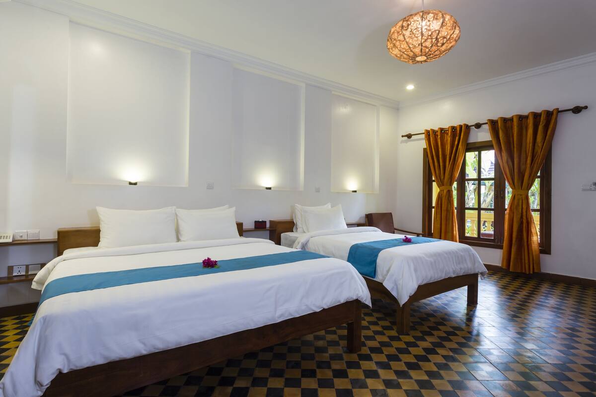 Une chambre d’hôtel avec deux lits, un sol à carreaux, une grande fenêtre avec des rideaux et des lampes fixées au mur.