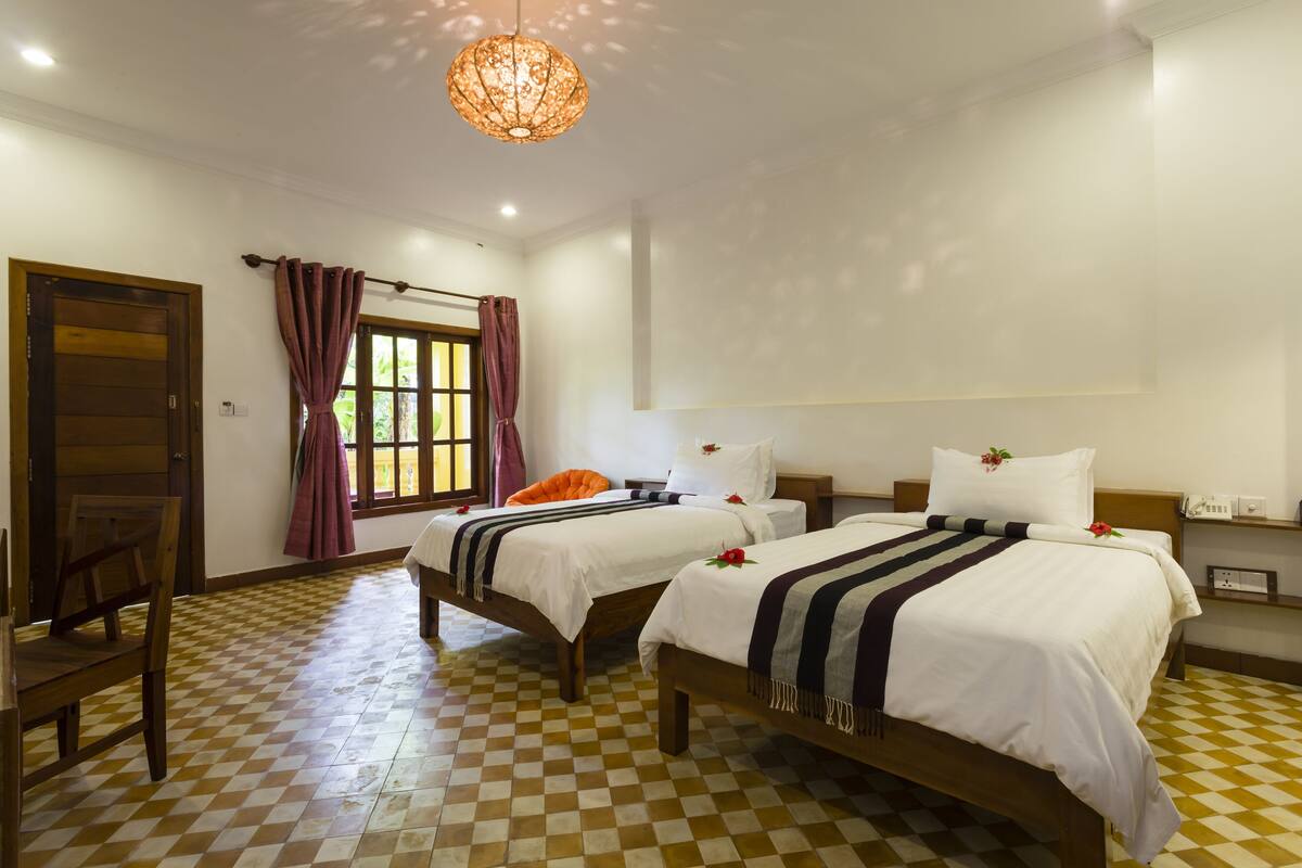 Une chambre d’hôtel avec deux lits, une chaise en bois, un sol à carreaux, une fenêtre avec des rideaux et un luminaire suspendu.