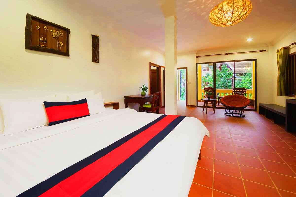 Une chambre d’hôtel avec un grand lit, un bureau avec une chaise, un balcon avec une chaise et une vue sur la verdure environnante.