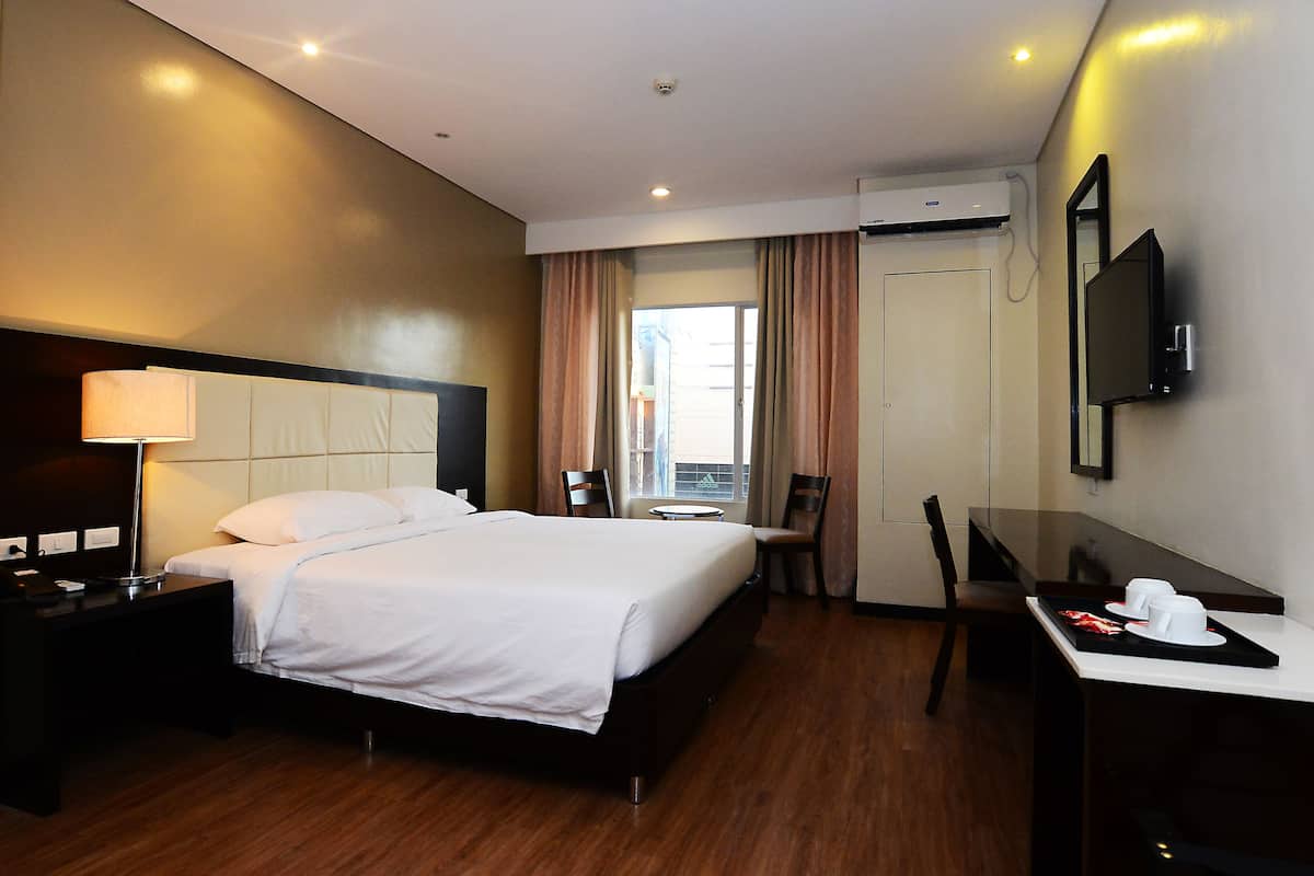 A hotel room with a bed, bedside table, lamp, TV, desk, chairs, and a window with curtains.