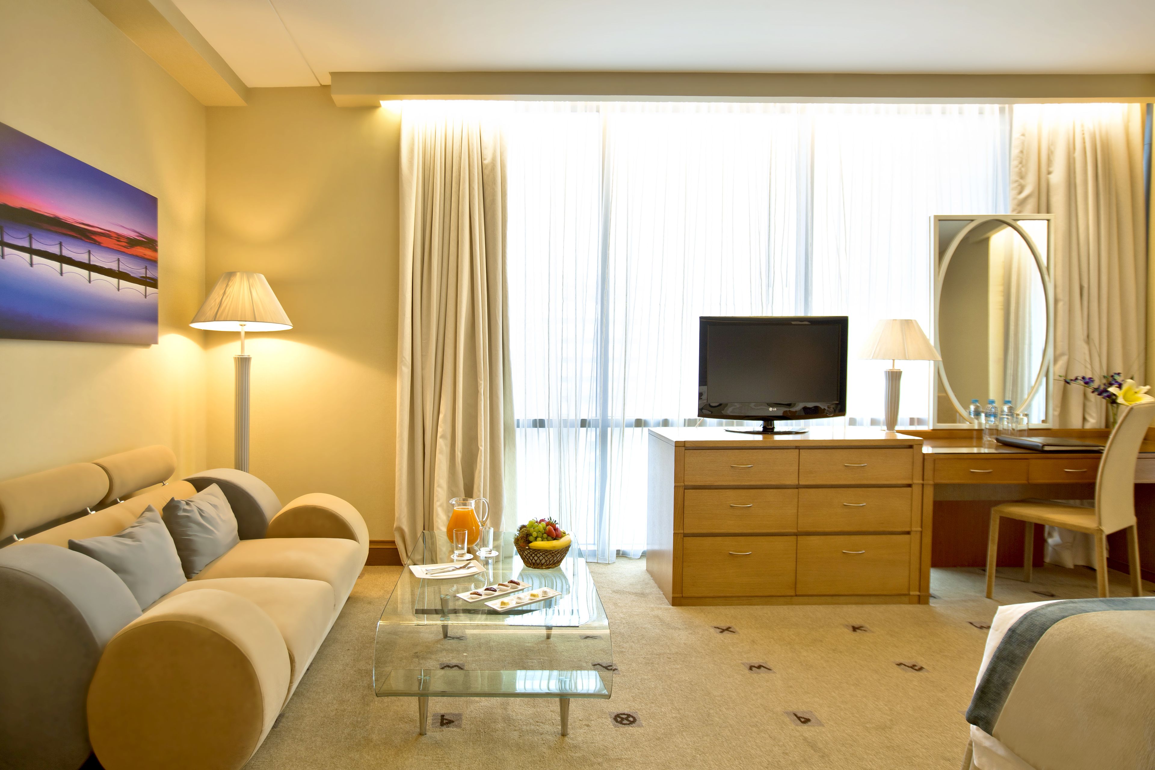 Hotelkamer met een bank, een glazen salontafel met een fruitschaal, een flatscreen televisie, een bureau met stoel, en een groot raam met gordijnen.