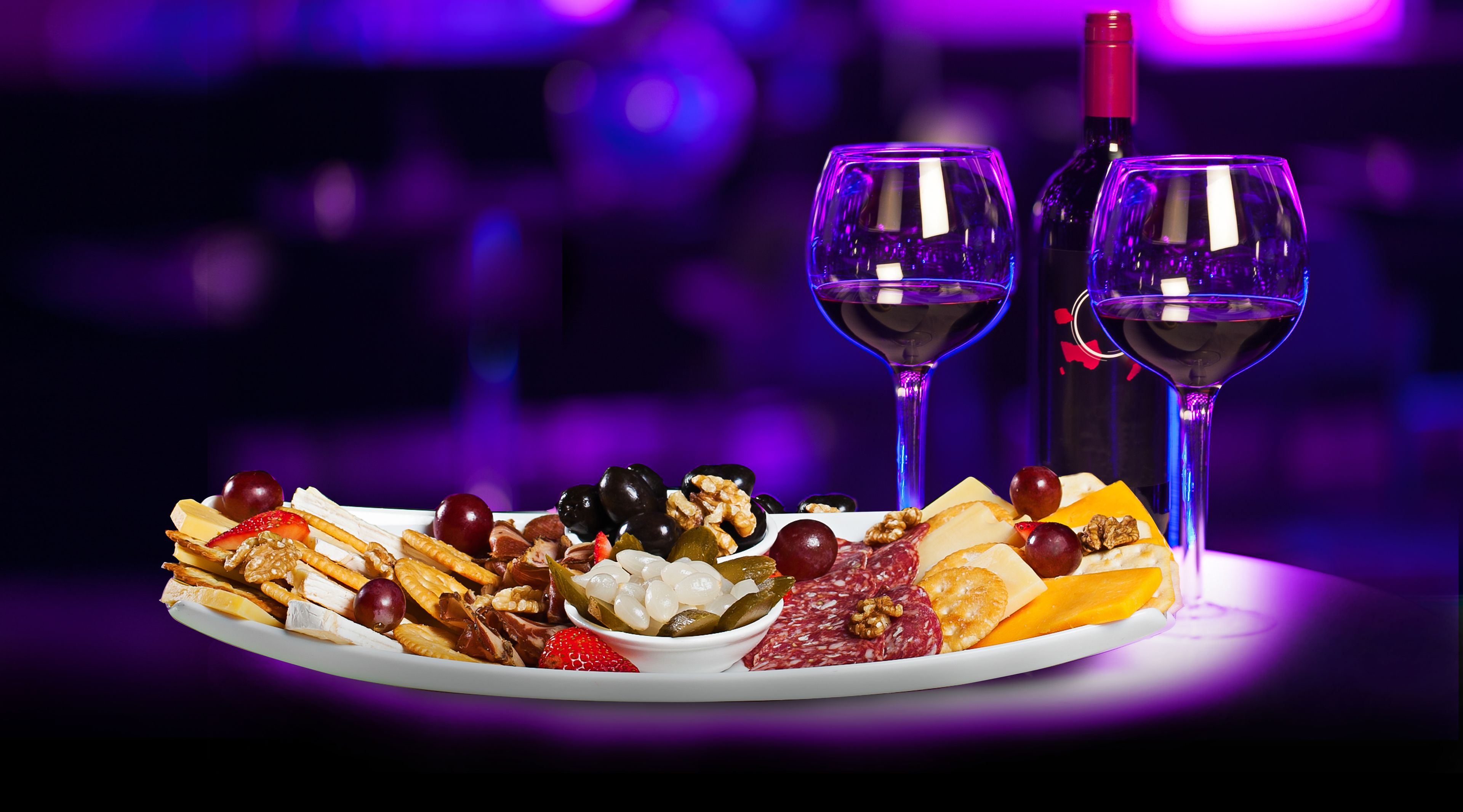 Un plateau garni d’un assortiment de fromages, de fruits et de noix, accompagné de deux verres à vin et d’une bouteille de vin.