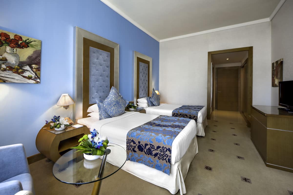 A hotel room with two beds, a glass table with a vase of flowers, a lamp, and a painting on the wall.