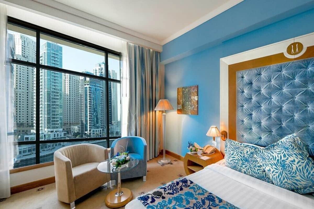 A hotel room with a large window, a bed with blue patterned pillows, a blue armchair, a small round table, and a nightstand with a lamp.