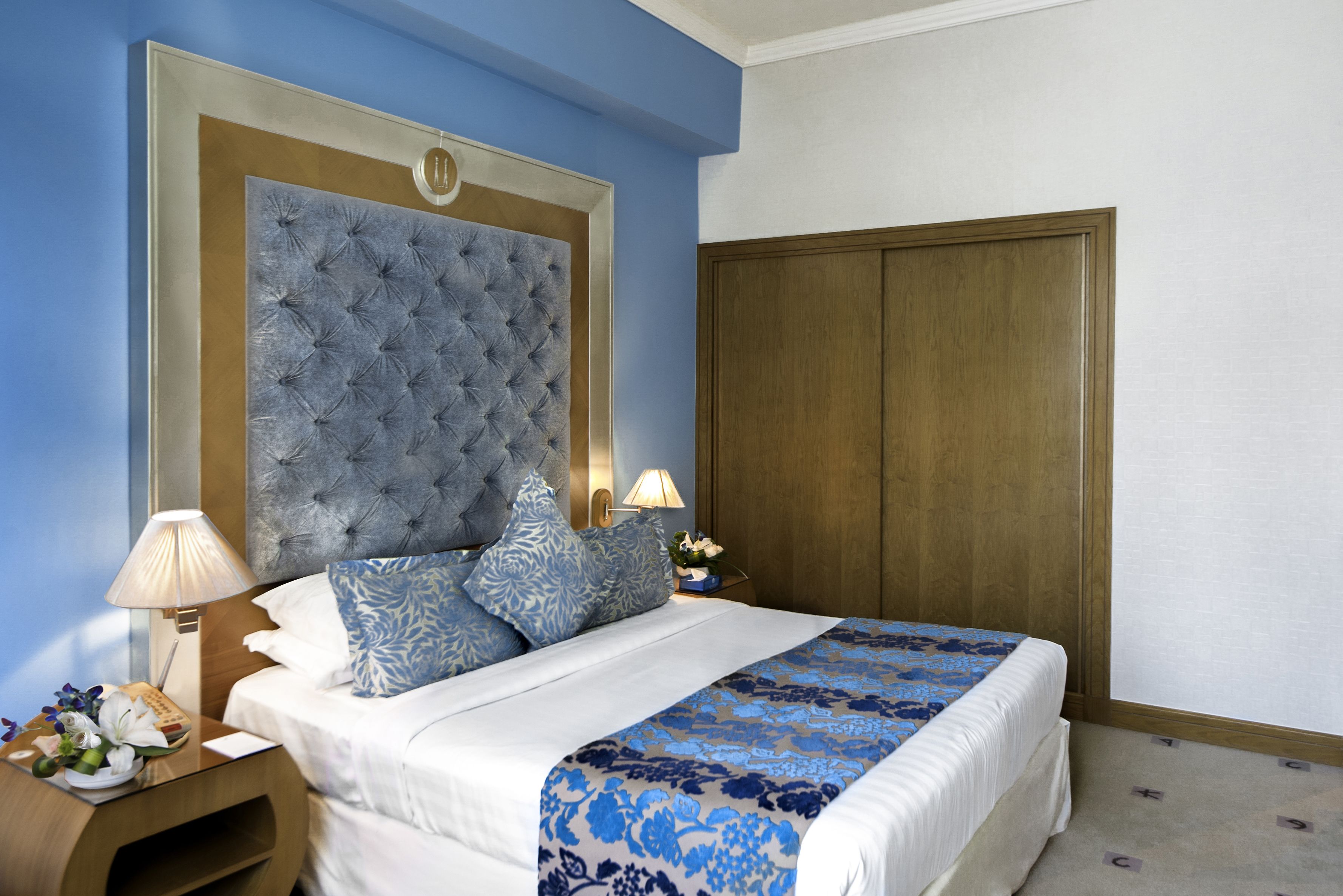 A hotel room with a large tufted headboard, a wooden wardrobe, a bedside table with a lamp, and a floral bedspread.