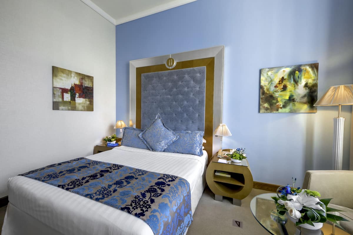 A hotel room with a large bed, a nightstand with a lamp, a glass table with flowers, and two paintings on the walls.