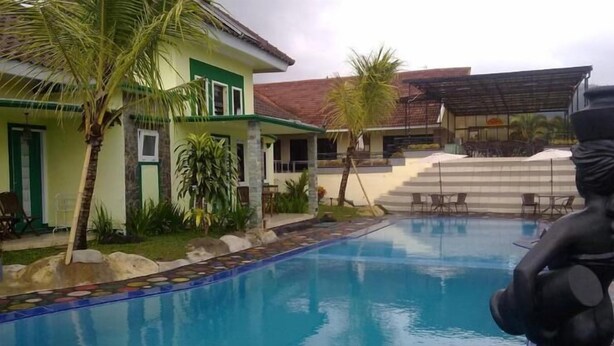 A swimming pool with a tiled edge, surrounded by a small garden with palm trees and a building with a green facade. - Seulawah Resort And Cafe (Batu)