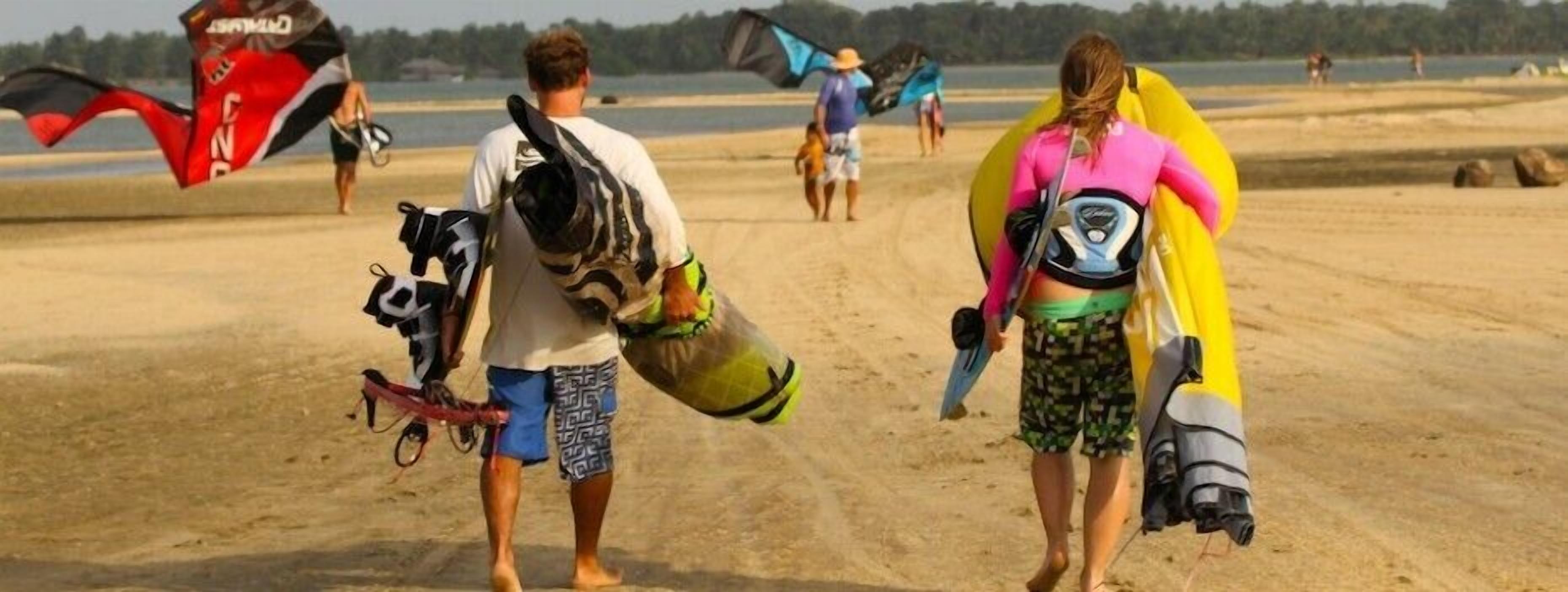 Duas pessoas caminham pela praia de areia carregando pranchas de surf e kitesurf.