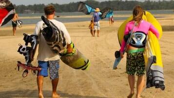 Duas pessoas caminham pela praia de areia carregando pranchas de surf e kitesurf.