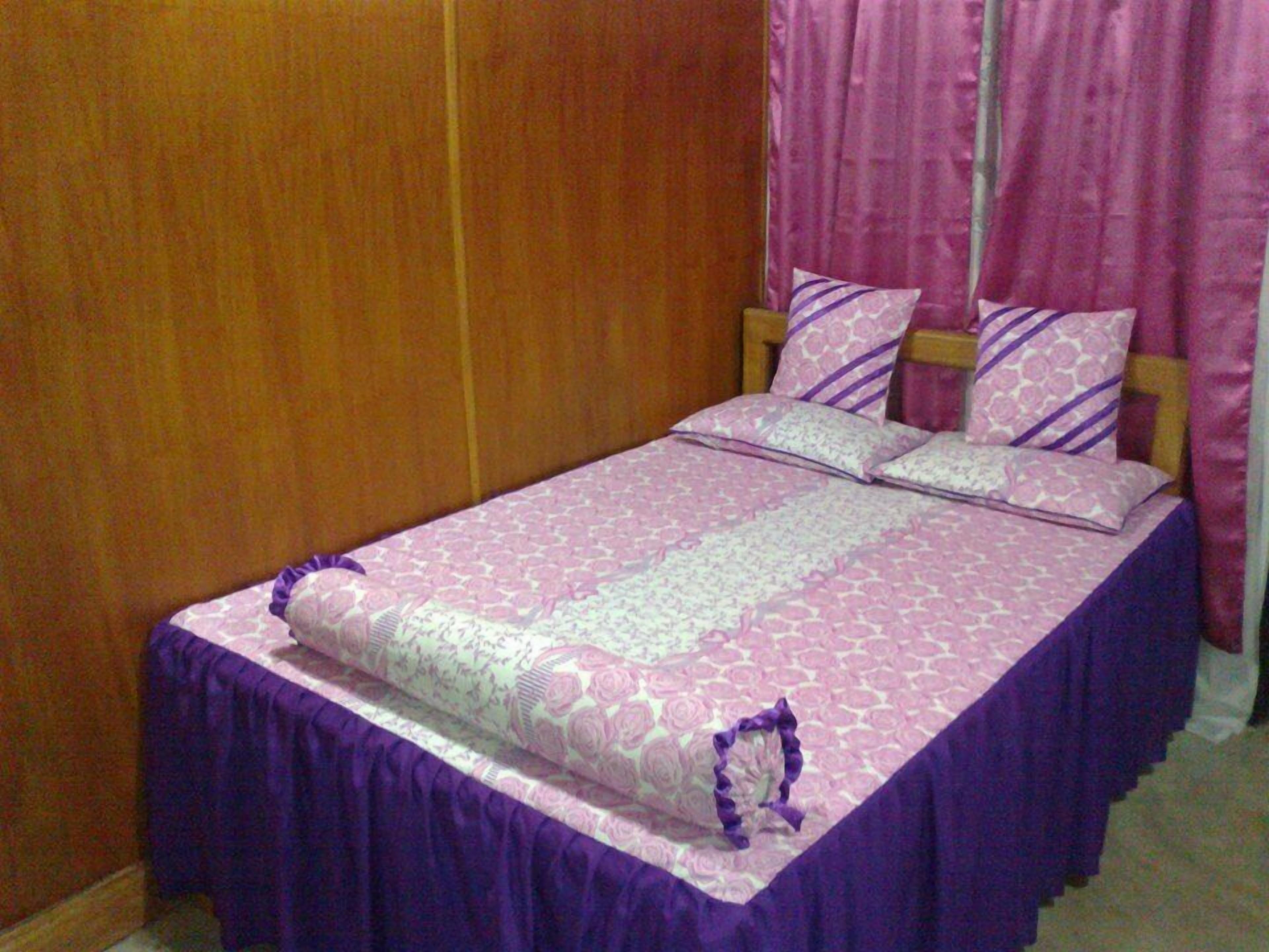 Un dormitorio con un armario de madera, una cama con colcha y almohadas moradas a juego, y cortinas rosas.