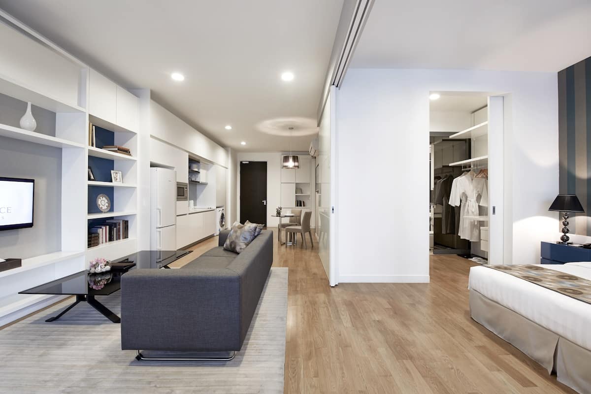 A modern living room with a grey sofa, a flat-screen TV, and a bookshelf. There is a dining area with a table and chairs, and a kitchen in the background.
