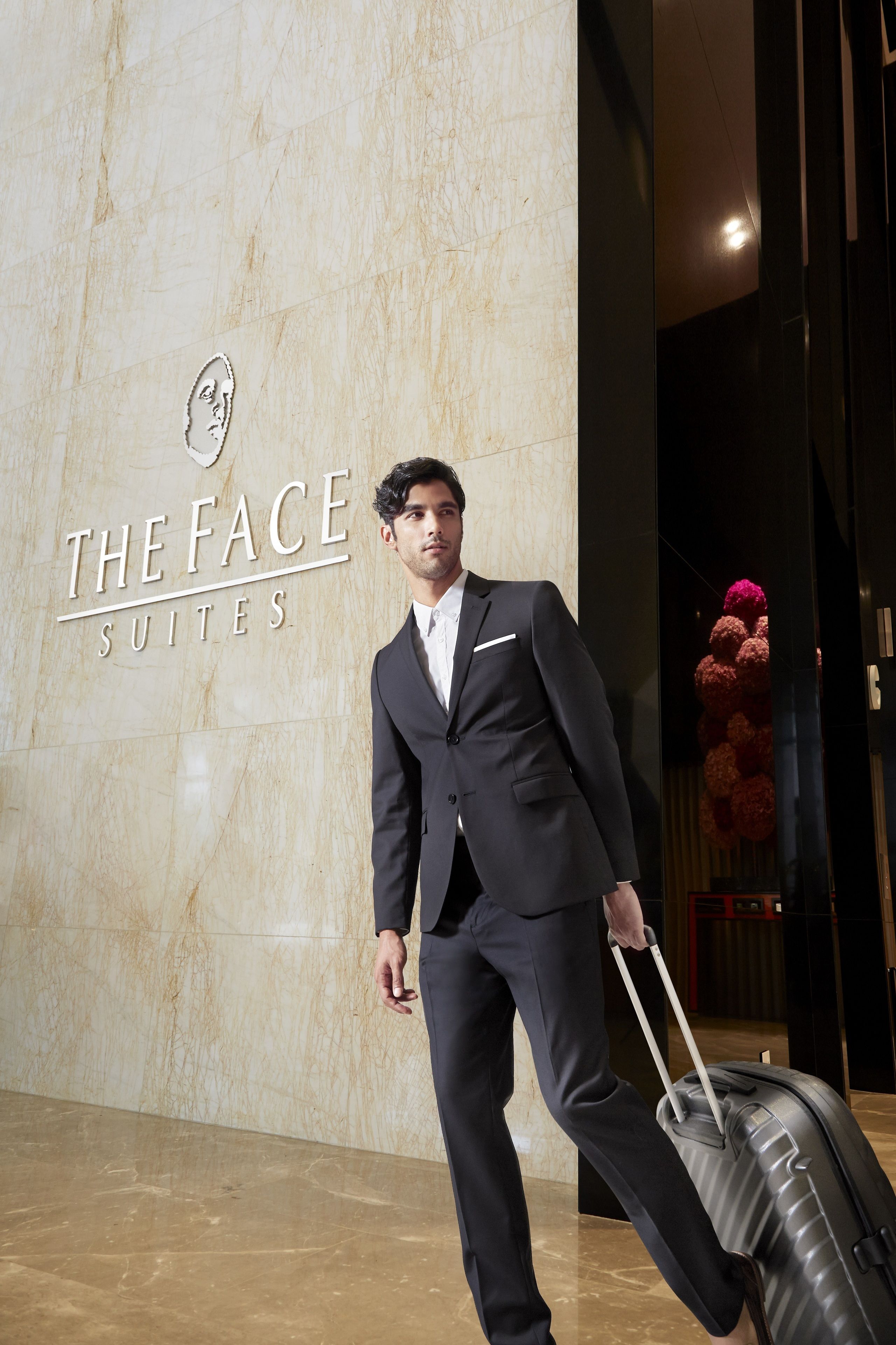 A man in a black suit with a white shirt and tie, pulling a black suitcase. The background features a marble wall with the text "THE FACE SUITES" and a decorative floral arrangement.