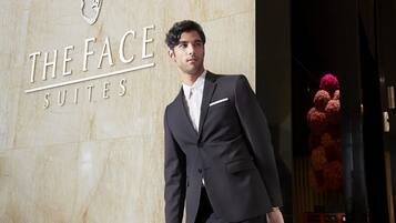 A man in a black suit with a white shirt and tie, pulling a black suitcase. The background features a marble wall with the text "THE FACE SUITES" and a decorative floral arrangement.