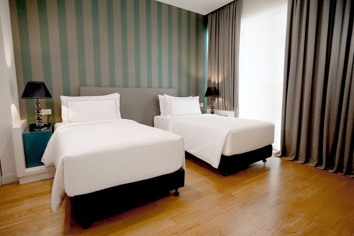 A hotel room with two beds, a nightstand with a lamp, and a view of the street through a window with curtains.