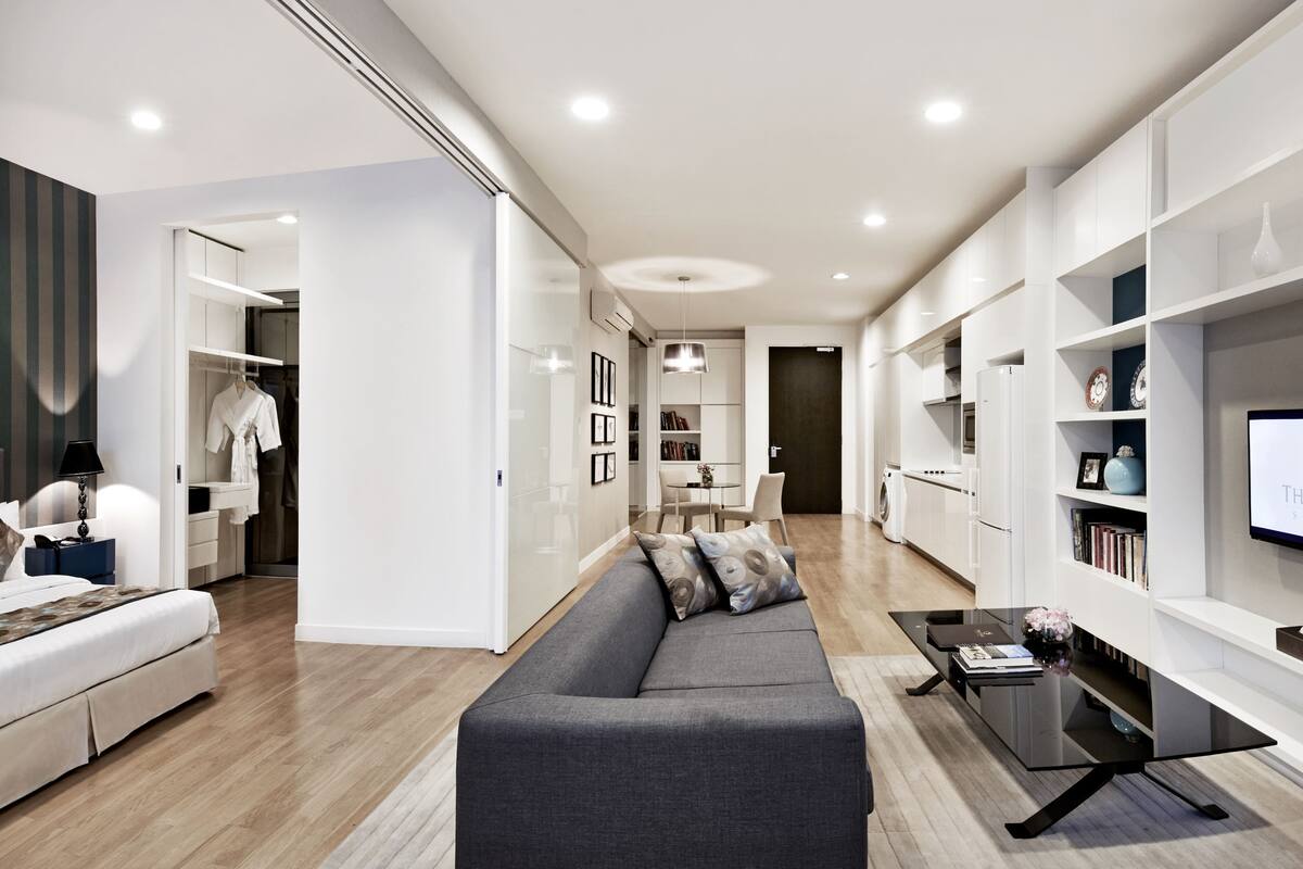 A modern living room with a grey sofa, a coffee table, and a TV mounted on the wall. There is a bookshelf with decorative items and a door leading to a laundry area.