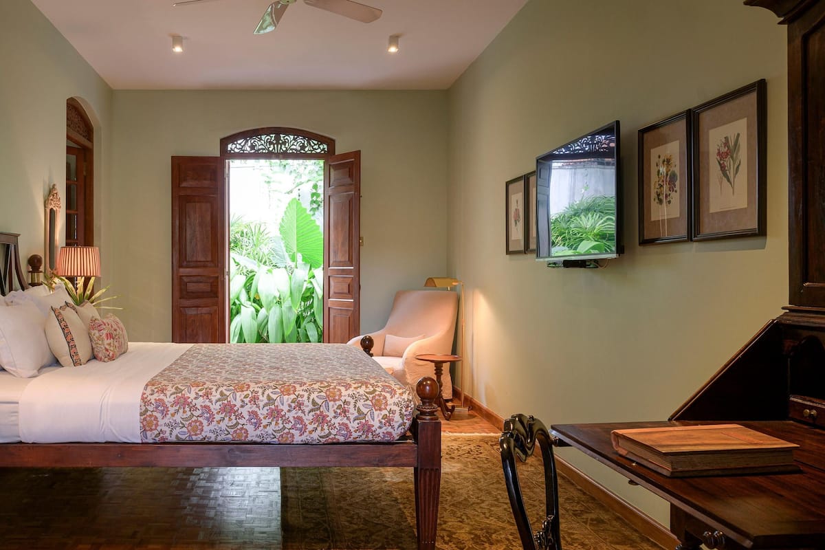 Un dormitorio con cama, mesita de noche, lámpara, silla, escritorio y un gran ventanal con vistas al verdor.