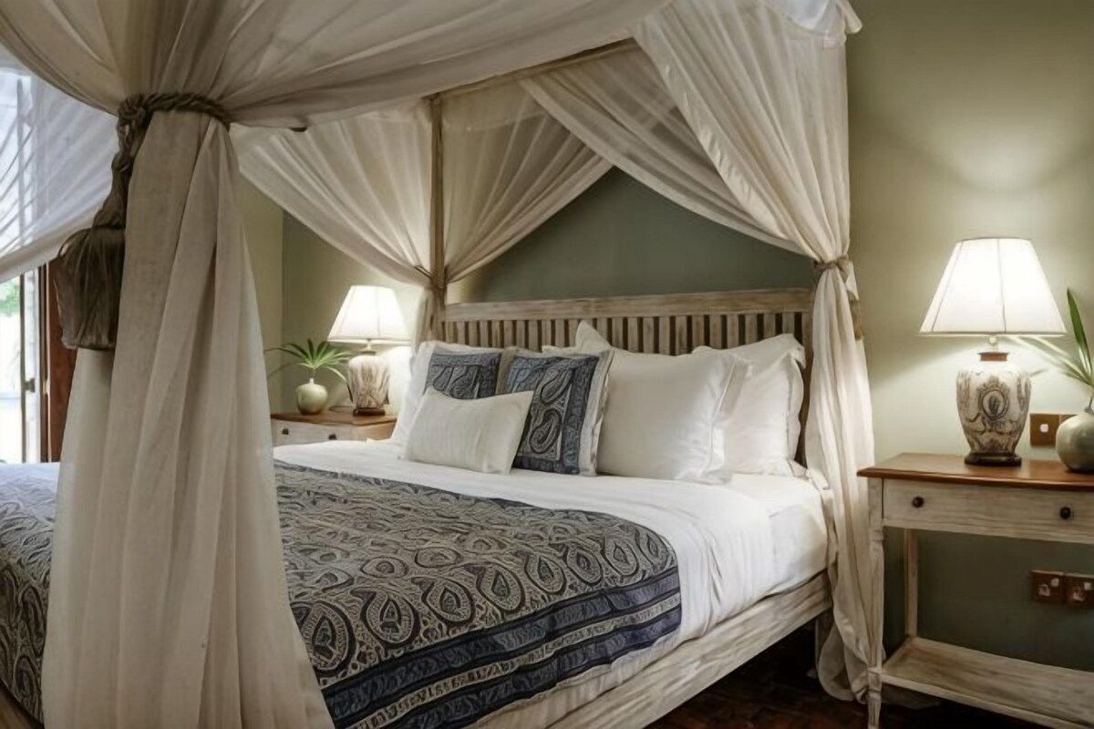 Uma cama com dossel e cortina, com edredom branco e azul estampado, duas mesas de cabeceira com luminárias e uma mesa de apoio com abajur.