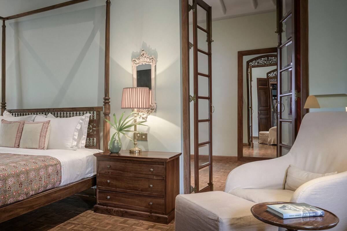 Una cama con dosel con un edredón floral, una mesita de noche de madera con cajones, un sillón blanco y una pequeña mesa redonda con un libro.