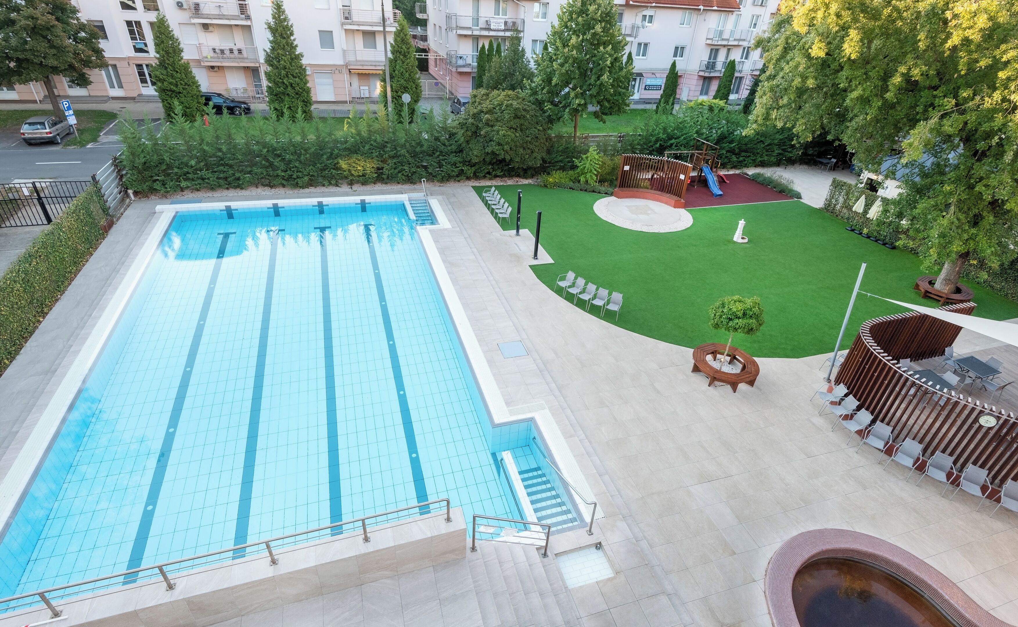 Ein Schwimmbad mit Fliesen umgeben, ein Kinderspielbereich und ein kleiner Garten mit Baum und Sitzgelegenheit.