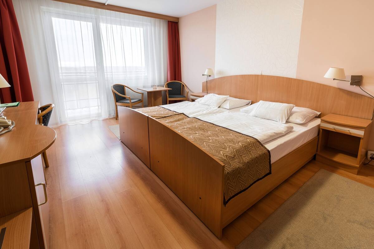 A hotel room with a large bed, a desk, a chair, a small table, and a window with curtains.