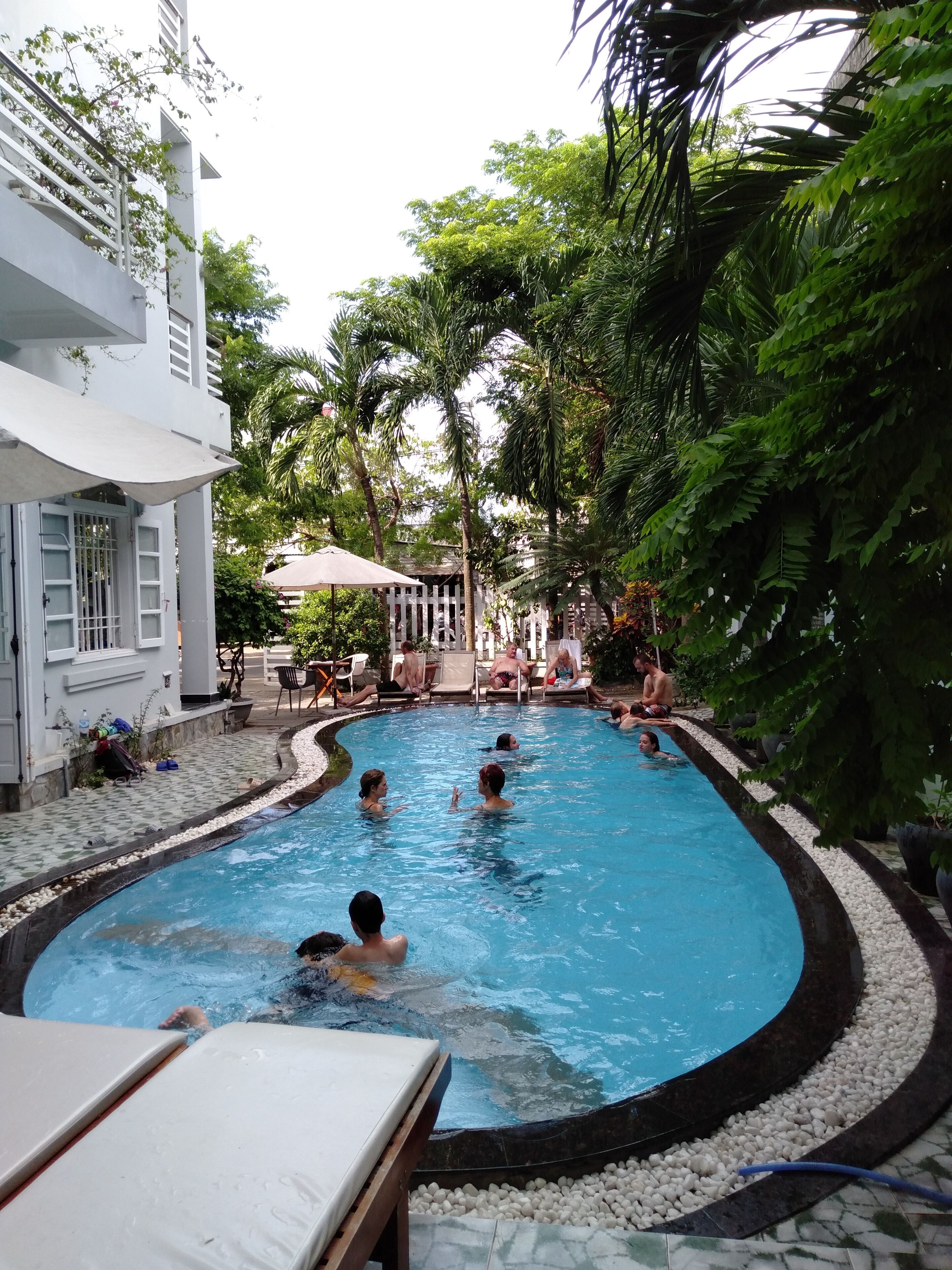A swimming pool area with people swimming and lounging, surrounded by lush greenery and a white building with a covered seating area.