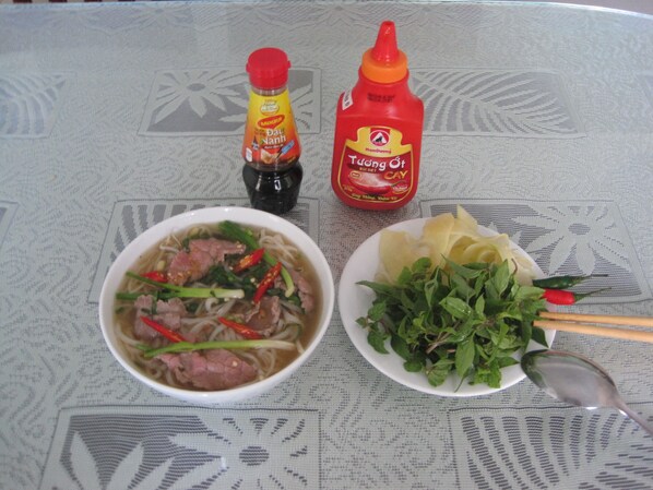 Una mesa con un tazón de sopa de fideos, un plato de vegetales verdes y dos botellas de salsas.
