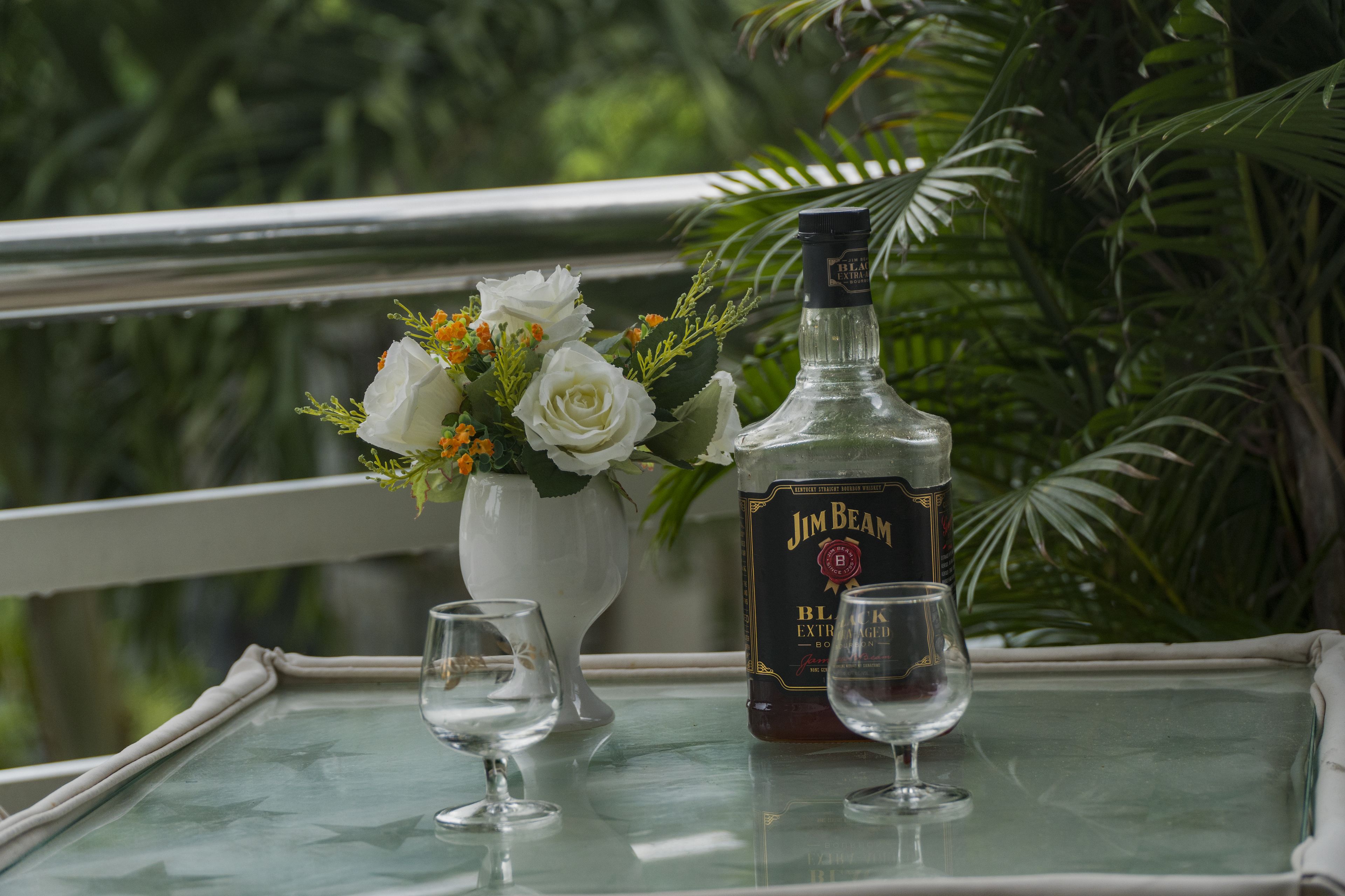 Uma mesa com uma garrafa de uísque Jim Beam, duas taças e um vaso com flores.