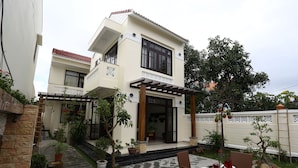 A two-story house with a patio area featuring a dining table set for four, surrounded by potted plants and a stone pathway. - Hoi An Cottage Villa (Da Nang)