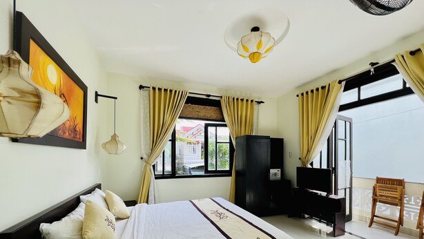 A hotel room with a bed, a TV, a wardrobe, and a balcony with a chair. - Hoi An Cottage Villa (Da Nang)