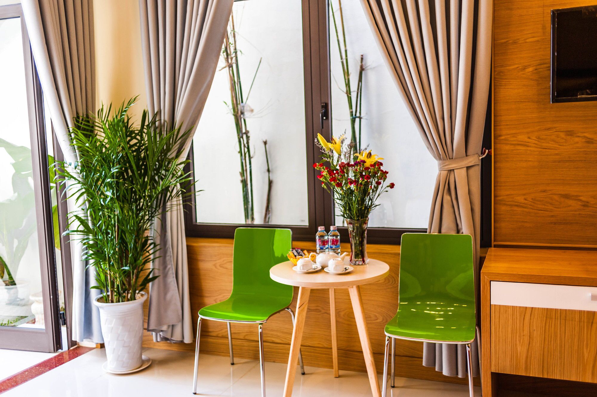 Habitación de hotel con dos sillas verdes, una pequeña mesa redonda con un jarrón de flores y una planta en maceta.
