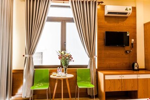 A room with two green chairs, a small round table with a vase of flowers, a wooden desk, and a flat-screen TV mounted on the wall. - Bien Dao Homestay Hoi An (Da Nang)