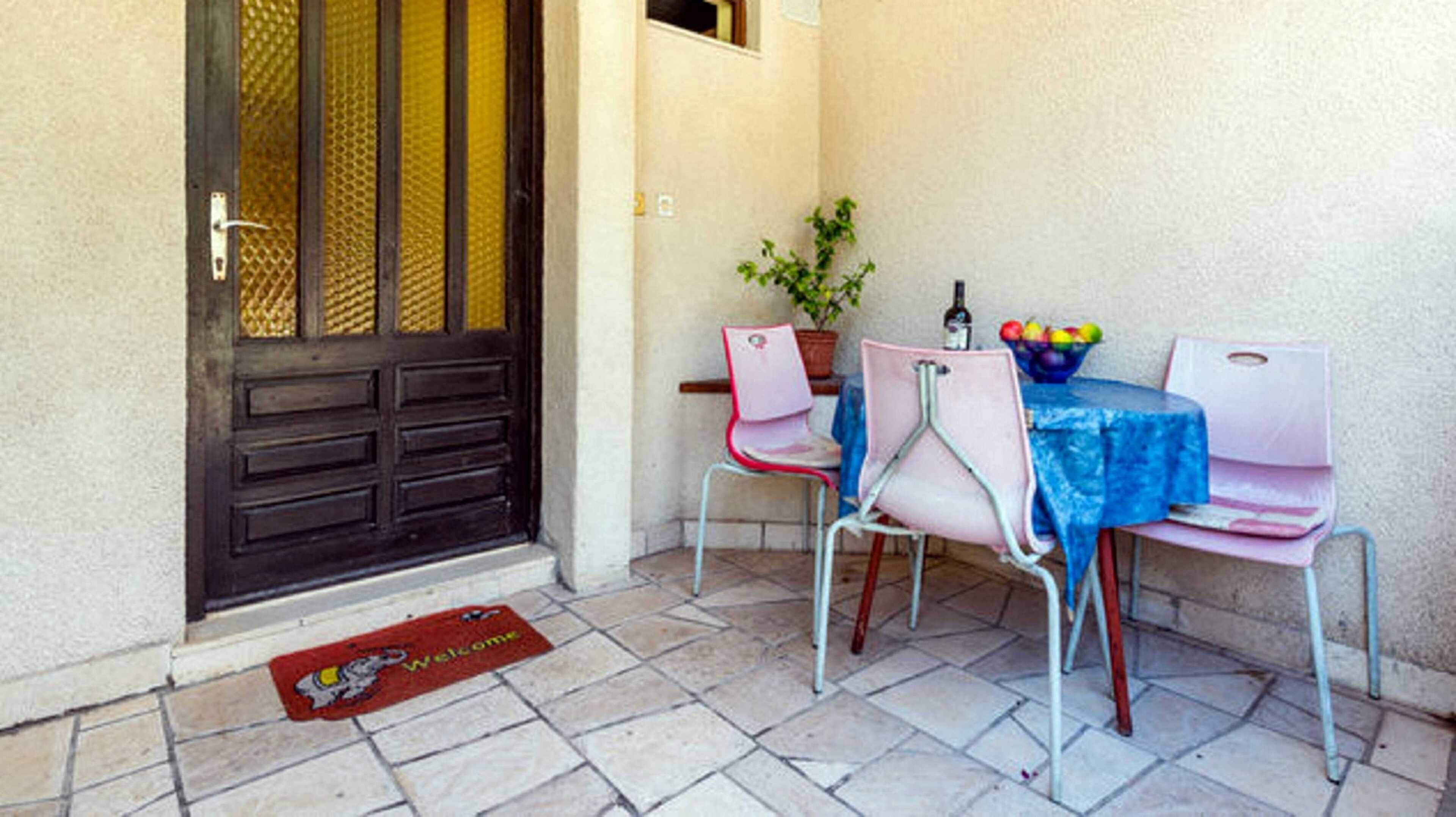 Een klein terras met een tafel en stoelen, een deur met hor, een bloempot en een welkomstmat.
