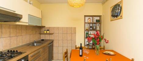 Une cuisine compacte équipée d’une cuisinière à gaz, d’un four et d’un four à micro-ondes. On y trouve une table à manger avec des chaises, un vase avec des fleurs et une bouteille de vin.